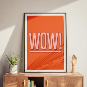 May include: A framed art print with the word "WOW!" in pink, set against a vibrant orange background. The print is displayed on a wooden cabinet, with a small potted plant and a wooden hand sculpture nearby.