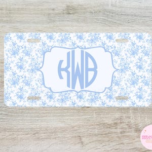 May include: A rectangular license plate featuring a blue floral pattern on a white background. The center has a light blue framed area with the initials "KWB" in a matching light blue font. The plate has four mounting holes.