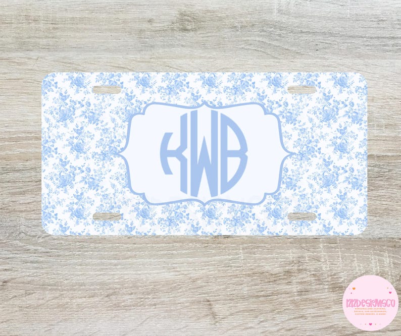May include: A rectangular license plate featuring a blue floral pattern on a white background. The center has a light blue framed area with the initials "KWB" in a matching light blue font. The plate has four mounting holes.