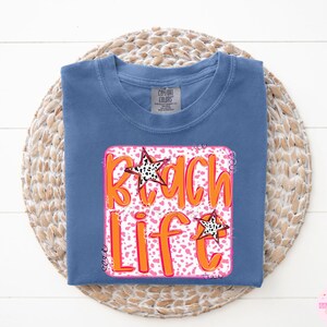 Puede incluir: Una camiseta azul con un gr&aacute;fico rosa y naranja que dice "Beach Life" con un dise&ntilde;o de estrella.