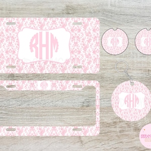 May include: A set of pink and white personalized car accessories featuring a damask pattern and the monogram "RHM". The set includes a license plate frame, two car coasters, and a key chain.