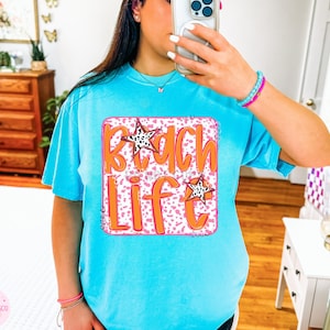 Puede incluir: Una camiseta azul turquesa con un gr&aacute;fico rosa y blanco que dice "Beach Life" en letras naranjas. El gr&aacute;fico tiene dos estrellas blancas y negras.
