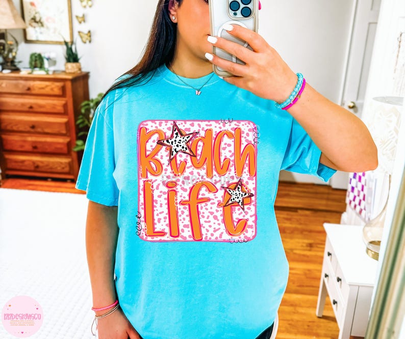Puede incluir: Una camiseta azul turquesa con un gr&aacute;fico rosa y blanco que dice "Beach Life" en letras naranjas. El gr&aacute;fico tiene dos estrellas blancas y negras.