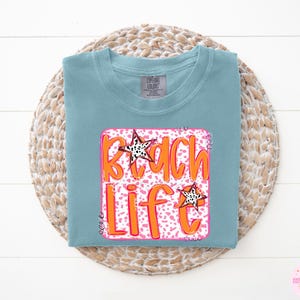 Puede incluir: Una camiseta azul claro con un gr&aacute;fico rosa y naranja que dice "Beach Life" con un dise&ntilde;o de estrella.