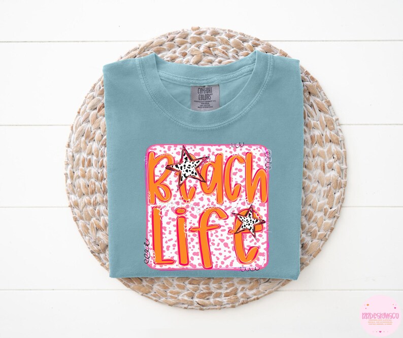 Puede incluir: Una camiseta azul claro con un gr&aacute;fico rosa y naranja que dice "Beach Life" con un dise&ntilde;o de estrella.