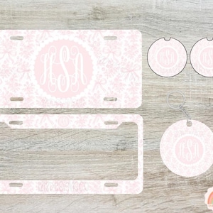 May include: A light pink license plate, license plate frame, two car coasters, and a keychain. All items feature a white floral pattern and a pink monogram. The license plate and frame have a rectangular shape, while the coasters are circular.