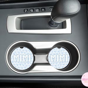 May include: Two car coasters with the name "MIMI" in white bubble letters. The coasters have a light blue snowflake pattern background. The coasters are in the car's cup holders.