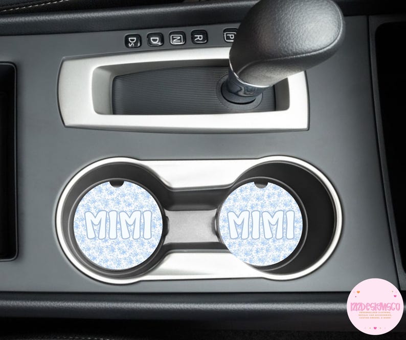 May include: Two car coasters with the name "MIMI" in white bubble letters. The coasters have a light blue snowflake pattern background. The coasters are in the car's cup holders.