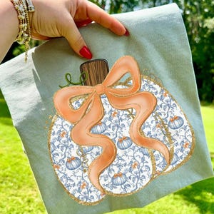 May include: A light green tea towel featuring a pumpkin design. The pumpkin has a white background with blue and orange floral patterns and blue pumpkins. An orange bow and gold trim complete the design.