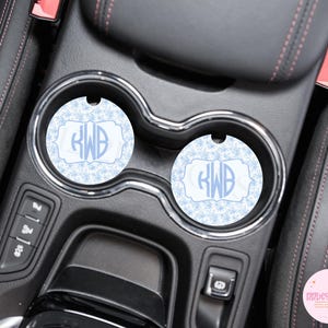 May include: Two light blue car coasters with a snowflake pattern and a monogram in a decorative frame. The coasters are placed in the cup holders of a car's center console.