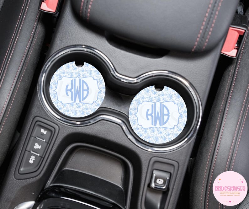 May include: Two light blue car coasters with a snowflake pattern and a monogram in a decorative frame. The coasters are placed in the cup holders of a car's center console.