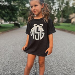 Puede incluir: Camiseta negra con un dise&ntilde;o de monograma blanco. El monograma presenta las letras "MSG" dentro de un dise&ntilde;o circular con un lazo en la parte superior. La camiseta la lleva una persona joven.