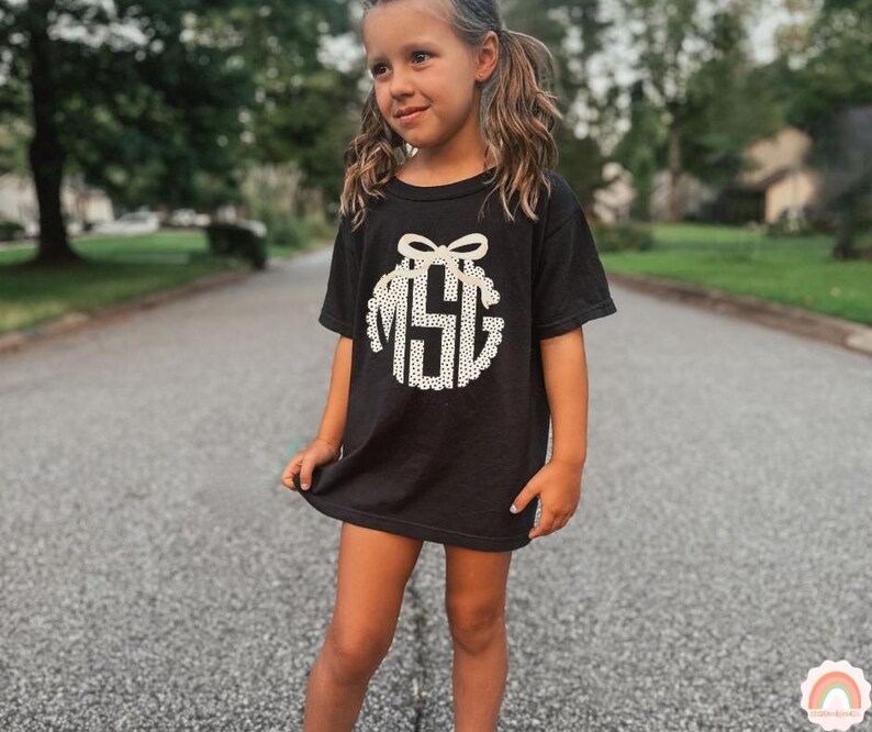 Puede incluir: Camiseta negra con un dise&ntilde;o de monograma blanco. El monograma presenta las letras "MSG" dentro de un dise&ntilde;o circular con un lazo en la parte superior. La camiseta la lleva una persona joven.