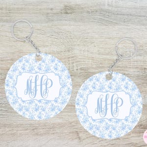 May include: Two round keychains with a light blue floral pattern and a white central design. Each keychain features the monogram "MHP" in a light blue script font, attached to a silver keyring and chain. The background is a light wood.