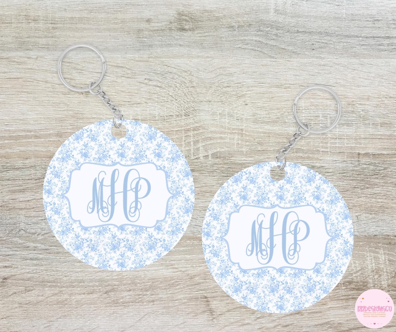 May include: Two round keychains with a light blue floral pattern and a white central design. Each keychain features the monogram "MHP" in a light blue script font, attached to a silver keyring and chain. The background is a light wood.