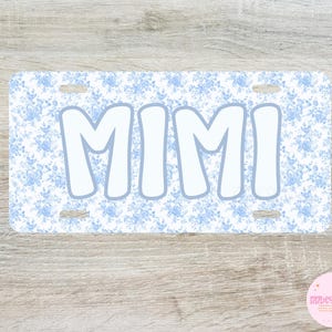 May include: A rectangular license plate with a light blue floral pattern background and the word "MIMI" in large, white, bubble letters. The plate has four mounting holes and a light blue border around the letters.