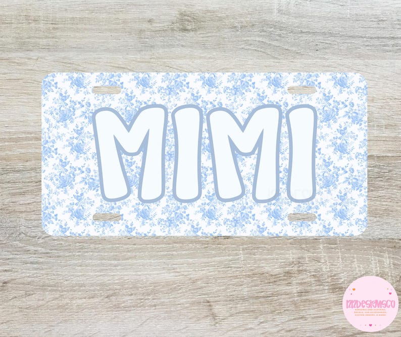 May include: A rectangular license plate with a light blue floral pattern background and the word "MIMI" in large, white, bubble letters. The plate has four mounting holes and a light blue border around the letters.
