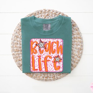 Puede incluir: Una camiseta verde azulada con un gr&aacute;fico rosa y naranja que dice "Beach Life" con una estrella blanca sobre un fondo rosa.