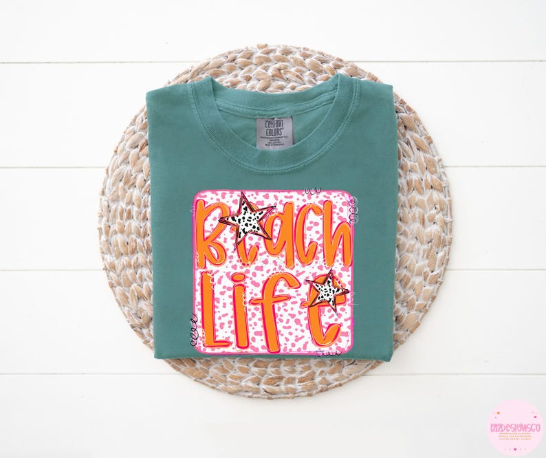 Puede incluir: Una camiseta verde azulada con un gr&aacute;fico rosa y naranja que dice "Beach Life" con una estrella blanca sobre un fondo rosa.