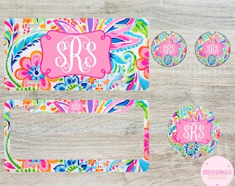 May include: A set of personalized car accessories featuring a colorful floral pattern with pink, blue, green, and white colors. The set includes a license plate frame, two round drink coasters, and a key chain. The monogram "SRS" is featured on each item.