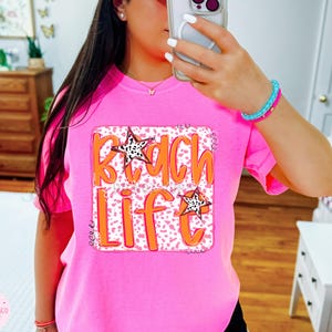 Puede incluir: Camiseta rosa con un dise&ntilde;o gr&aacute;fico que dice "Beach Life" en letras naranjas sobre fondo blanco y estrellas blancas y negras. El gr&aacute;fico est&aacute; rodeado por un borde blanco.