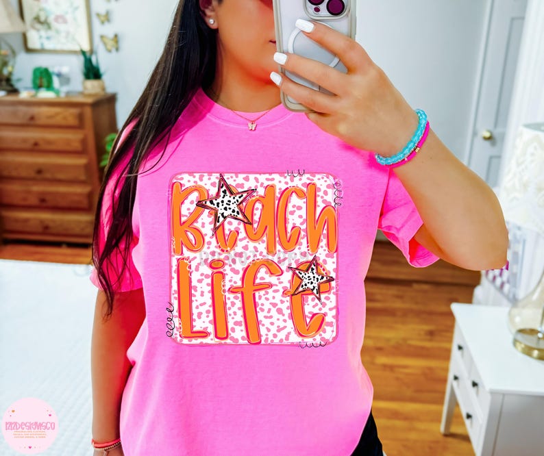 Puede incluir: Camiseta rosa con un dise&ntilde;o gr&aacute;fico que dice "Beach Life" en letras naranjas sobre fondo blanco y estrellas blancas y negras. El gr&aacute;fico est&aacute; rodeado por un borde blanco.