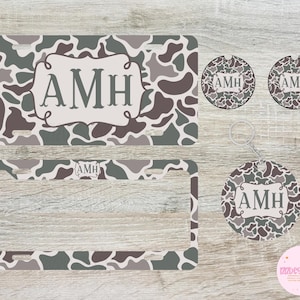 May include: A set of personalized camo-patterned car accessories featuring the initials "AMH". The set includes a license plate frame, two car coasters, and a key chain.