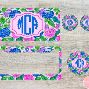 May include: A rectangular license plate, a license plate frame, two circular car coasters, and a circular keychain, all featuring a vibrant floral pattern of blue and pink hydrangeas with green leaves and a central pink-bordered label with the blue monogram MCA.