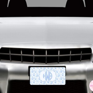 May include: A light blue and white license plate with a snowflake pattern and a monogram. The license plate is rectangular and is mounted on the front of a silver car. The monogram includes the letters "RWB".