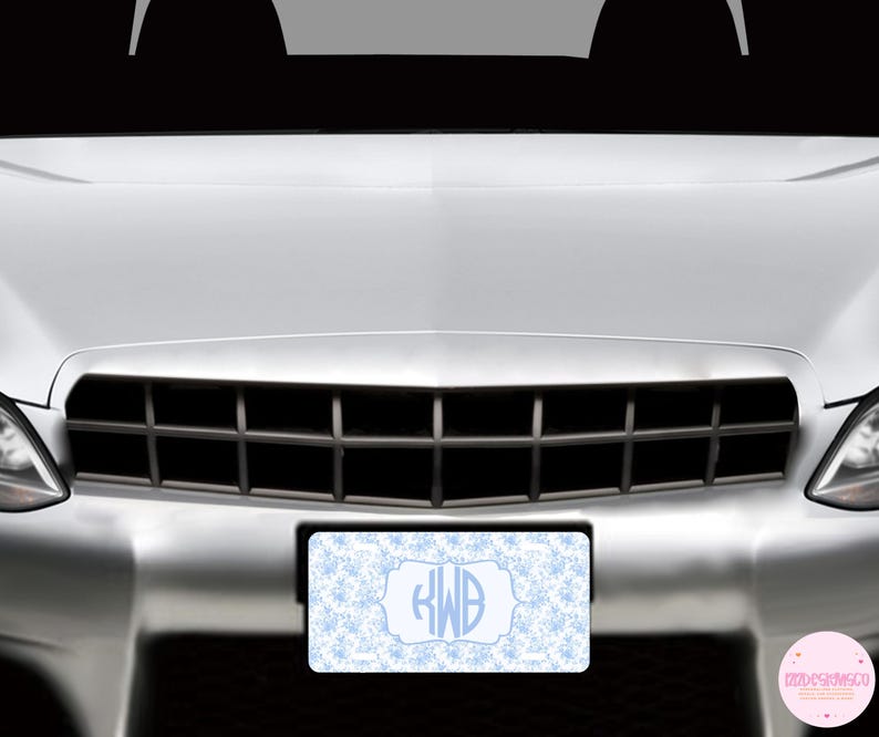 May include: A light blue and white license plate with a snowflake pattern and a monogram. The license plate is rectangular and is mounted on the front of a silver car. The monogram includes the letters "RWB".