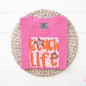 Puede incluir: Camiseta rosa con un estampado gr&aacute;fico de las palabras "Beach Life" en naranja sobre un fondo de estampado de leopardo rosa y blanco. El gr&aacute;fico tambi&eacute;n incluye dos estrellas doradas.