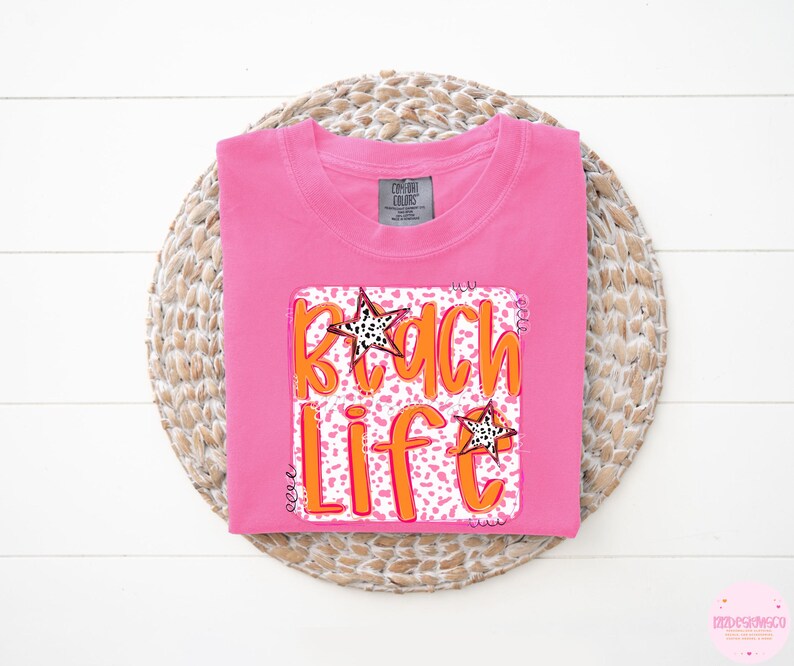 Puede incluir: Camiseta rosa con un estampado gr&aacute;fico de las palabras "Beach Life" en naranja sobre un fondo de estampado de leopardo rosa y blanco. El gr&aacute;fico tambi&eacute;n incluye dos estrellas doradas.