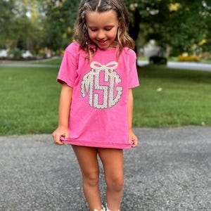 Puede incluir: Una camiseta rosa con un dise&ntilde;o de monograma y un lazo en la parte superior. El monograma tiene un patr&oacute;n de lunares blancos. La camiseta est&aacute; siendo sostenida por una persona.