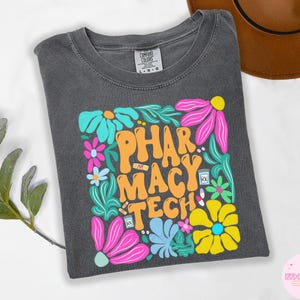 Puede incluir: Camiseta gris oscuro con la frase "PHARMACY TECH" en naranja, rodeada de flores coloridas y gráficos relacionados con la farmacia. La camiseta está hecha de un material suave y es adecuada para el uso diario.