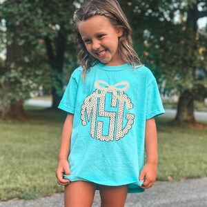 Puede incluir: Una camiseta turquesa con un dise&ntilde;o de monograma y un lazo. El monograma est&aacute; en un c&iacute;rculo con un borde festoneado y un estampado de lunares. La camiseta tiene mangas cortas.