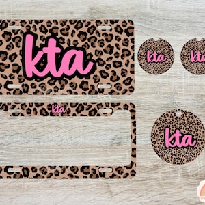May include: A set of car accessories with a leopard print design. Includes a license plate, license plate frame, two car coasters, and a keychain. Each item features the letters "kta" in pink.