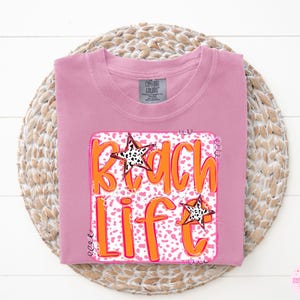 Puede incluir: Camiseta rosa con un estampado gr&aacute;fico de un dise&ntilde;o "Beach Life" rosa y naranja con estrellas blancas y negras.