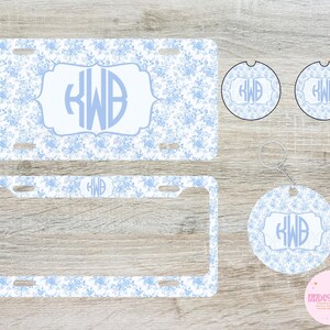 May include: A set of car accessories featuring a light blue floral pattern. The set includes a license plate, license plate frame, two car coasters, and a keychain, all with the initials "KWB" in a decorative frame.