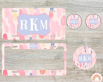 May include: A set of car accessories with a watercolor design. Includes a license plate, license plate frame, two car coasters, and a keychain. The items feature a pink, blue, and yellow brushstroke pattern and the initials "RKM".