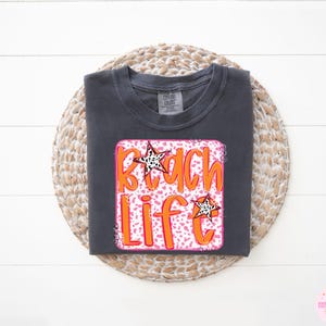 Puede incluir: Una camiseta gris oscura con un gr&aacute;fico rosa y naranja que dice "Beach Life" con un dise&ntilde;o de estrella.