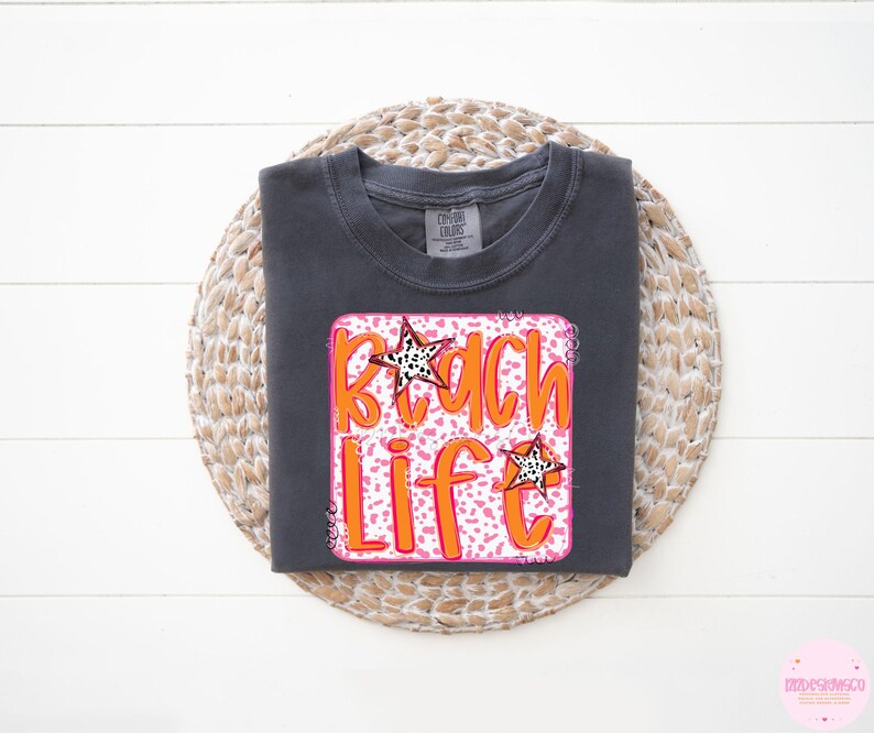 Puede incluir: Una camiseta gris oscura con un gr&aacute;fico rosa y naranja que dice "Beach Life" con un dise&ntilde;o de estrella.