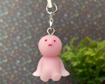 May include: A pink, doll-like charm with a small body, arms, and head. The charm has a pink string and a silver clasp. The background is a colorful, speckled pattern.