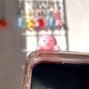 Puede incluir: Un charm de tel&eacute;fono rosa con forma de pulpo y expresi&oacute;n de sorpresa se encuentra sobre una funda transparente para tel&eacute;fono. El charm tiene peque&ntilde;os brazos y una cabeza redonda con ojos y boca rosas. La funda del tel&eacute;fono es transparente con un tinte rosa.