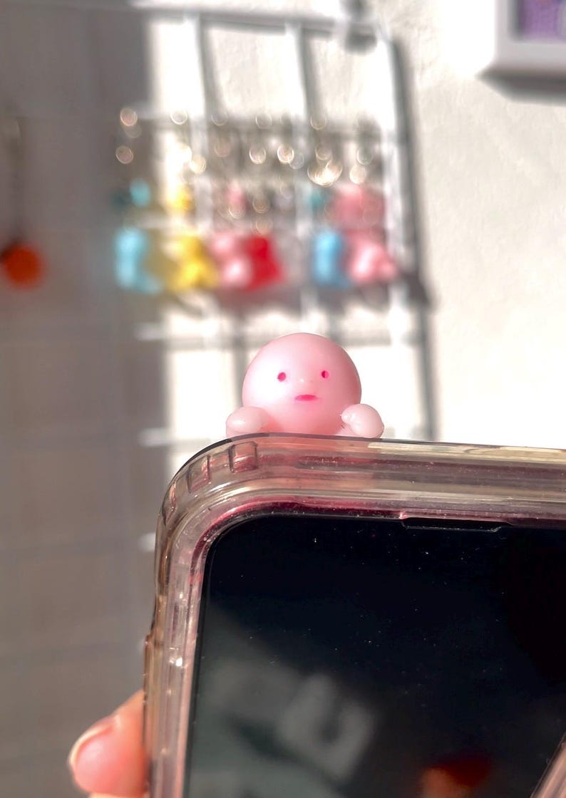 Puede incluir: Un charm de tel&eacute;fono rosa con forma de pulpo y expresi&oacute;n de sorpresa se encuentra sobre una funda transparente para tel&eacute;fono. El charm tiene peque&ntilde;os brazos y una cabeza redonda con ojos y boca rosas. La funda del tel&eacute;fono es transparente con un tinte rosa.