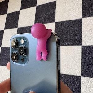 Puede incluir: Una figura rosa estilizada de una persona agarrada al lateral de un tel&eacute;fono inteligente azul. La figura es de color s&oacute;lido con una cabeza redondeada y un brazo extendido. El tel&eacute;fono tiene m&uacute;ltiples lentes de c&aacute;mara.
