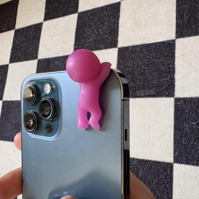 Puede incluir: Una figura rosa estilizada de una persona agarrada al lateral de un tel&eacute;fono inteligente azul. La figura es de color s&oacute;lido con una cabeza redondeada y un brazo extendido. El tel&eacute;fono tiene m&uacute;ltiples lentes de c&aacute;mara.