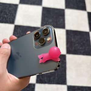 Puede incluir: Un accesorio de tel&eacute;fono de silicona rosa con forma de persona peque&ntilde;a est&aacute; adherido a la parte trasera de un smartphone gris oscuro. El tel&eacute;fono tiene tres lentes de c&aacute;mara y un marco plateado. El fondo es un patr&oacute;n de cuadros blanco y negro.