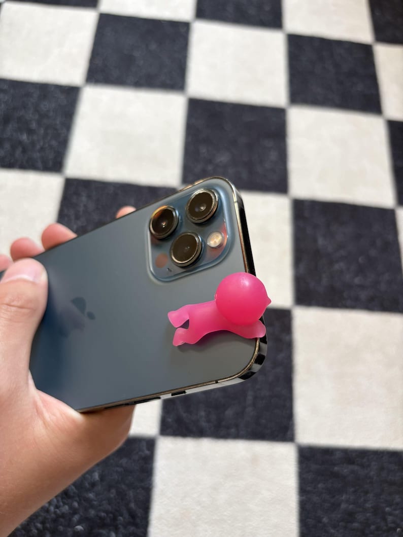 Puede incluir: Un accesorio de tel&eacute;fono de silicona rosa con forma de persona peque&ntilde;a est&aacute; adherido a la parte trasera de un smartphone gris oscuro. El tel&eacute;fono tiene tres lentes de c&aacute;mara y un marco plateado. El fondo es un patr&oacute;n de cuadros blanco y negro.