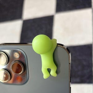 Puede incluir: Un colgante para tel&eacute;fono verde lima con forma de una peque&ntilde;a persona agarrada al borde de un smartphone gris. El tel&eacute;fono tiene m&uacute;ltiples lentes de c&aacute;mara. El fondo es un patr&oacute;n de cuadros blancos y negros.