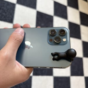 Puede incluir: Un smartphone de color verde azulado con una figura negra estilizada adjunta. El tel&eacute;fono tiene una c&aacute;mara de triple lente y el logotipo de Apple. La figura es una peque&ntilde;a forma humana negra abstracta con una cabeza redondeada.